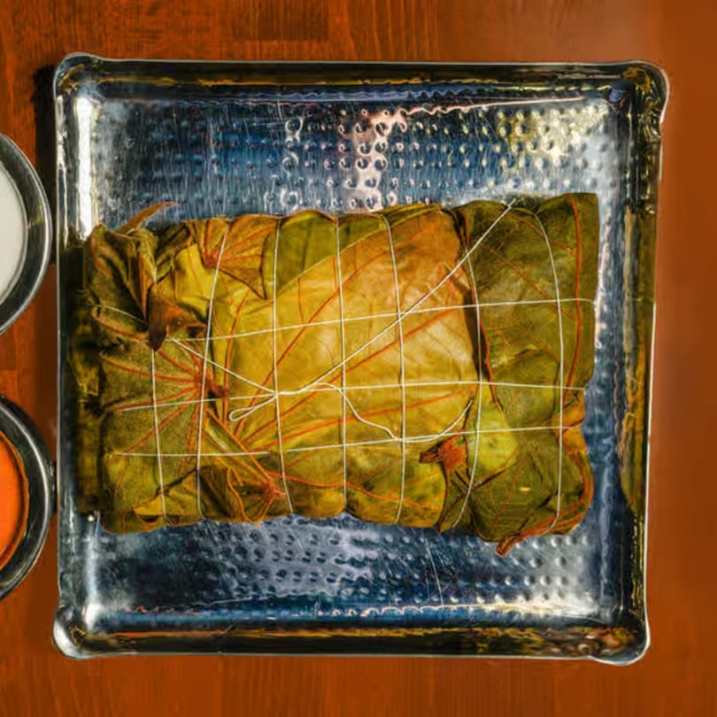 Indian dish wrapped in green leaves and tied with string, served on a silver tray.